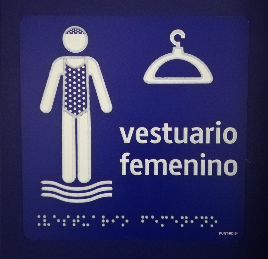 Señal vestuario femenino fondo azul con pictograma blanco de mujer en bañador y percha. También braille en blanco